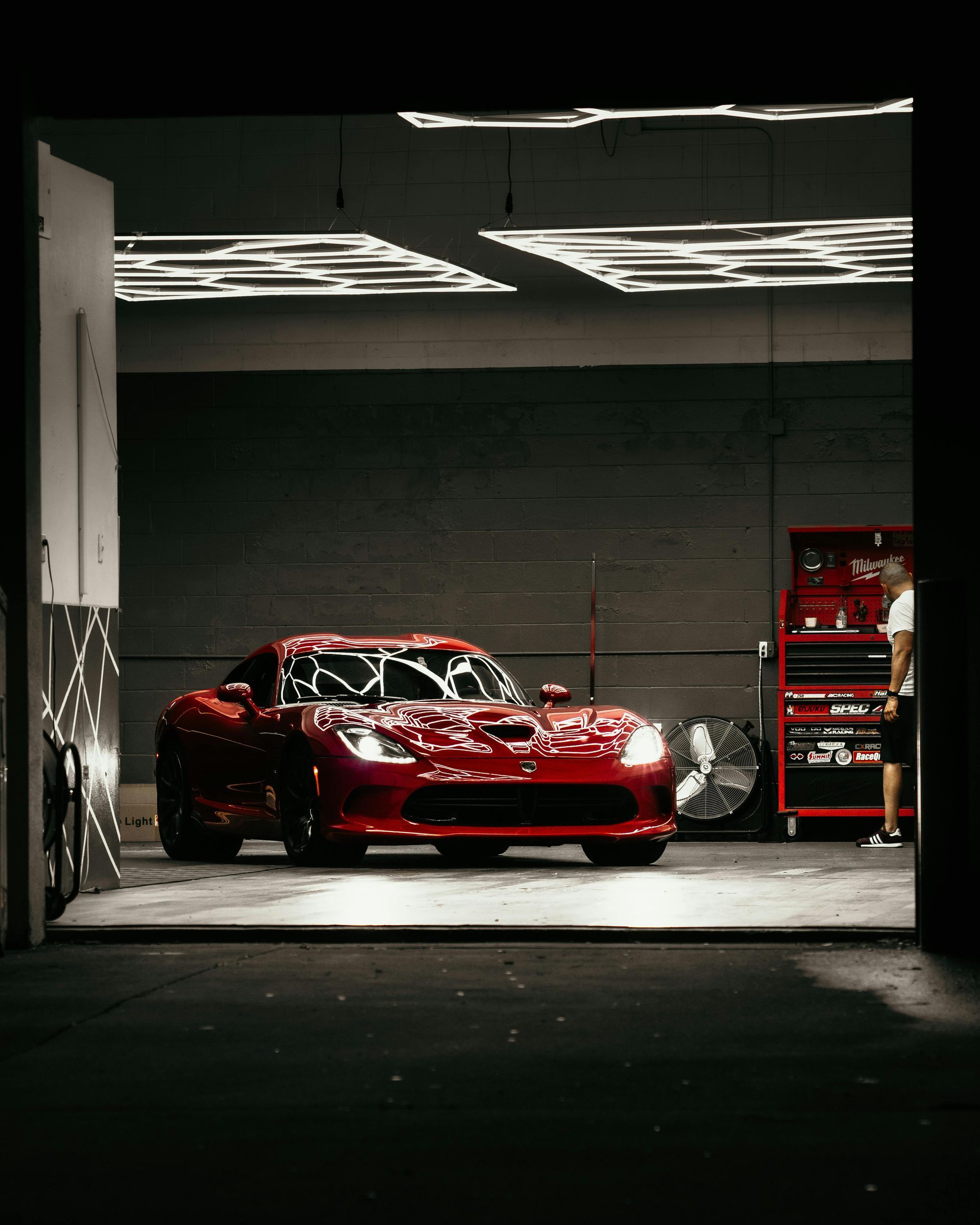 Red sports car in garage, lit by overhead lights. Person in background near tools.