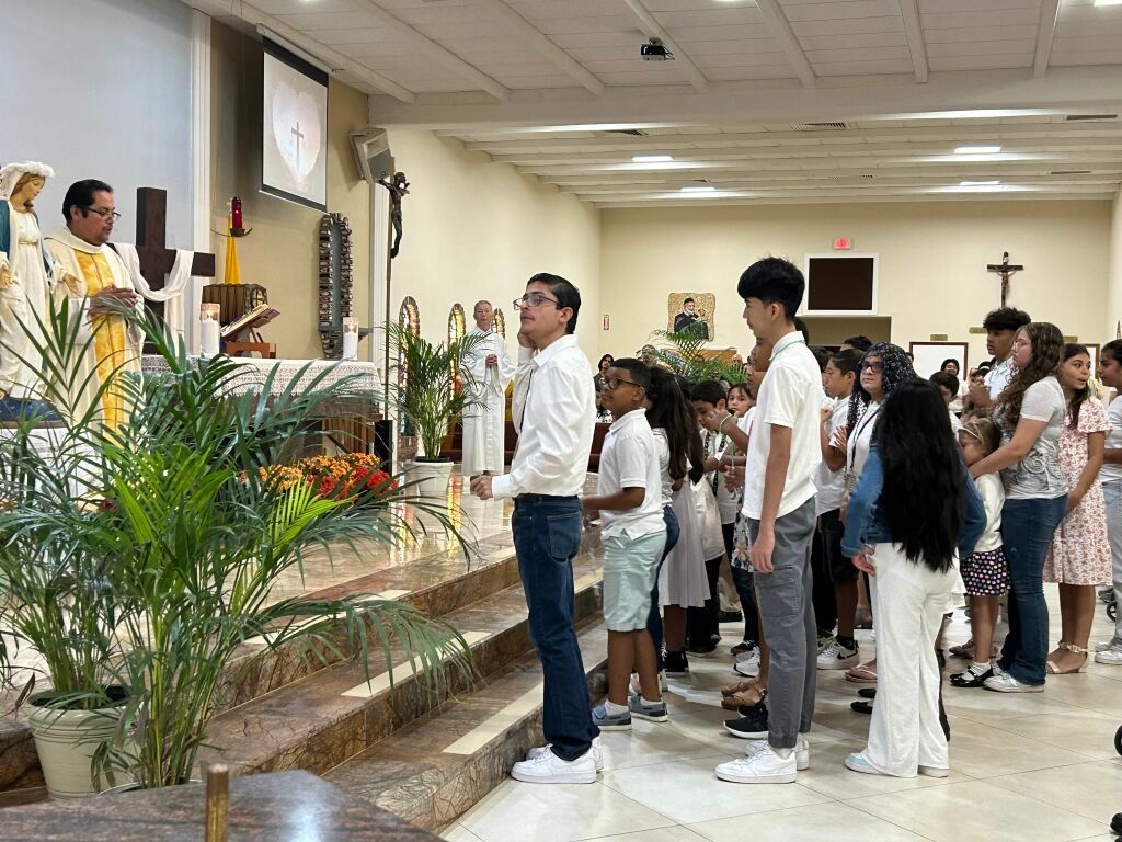 People standing in a church aisle during a service, facing the altar and flowers.