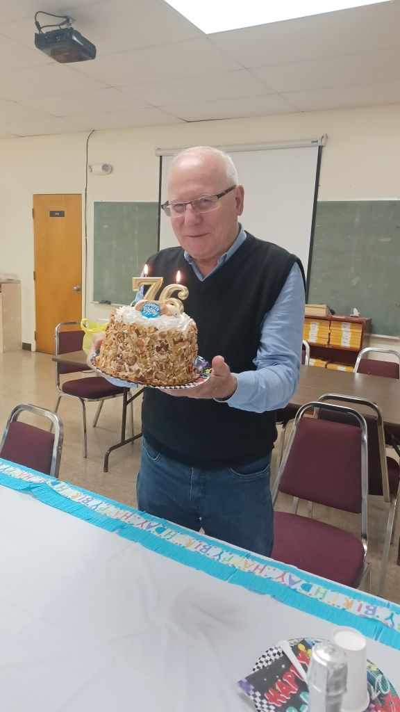 Man holding a decorated cake with “72” candles in a classroom party setting