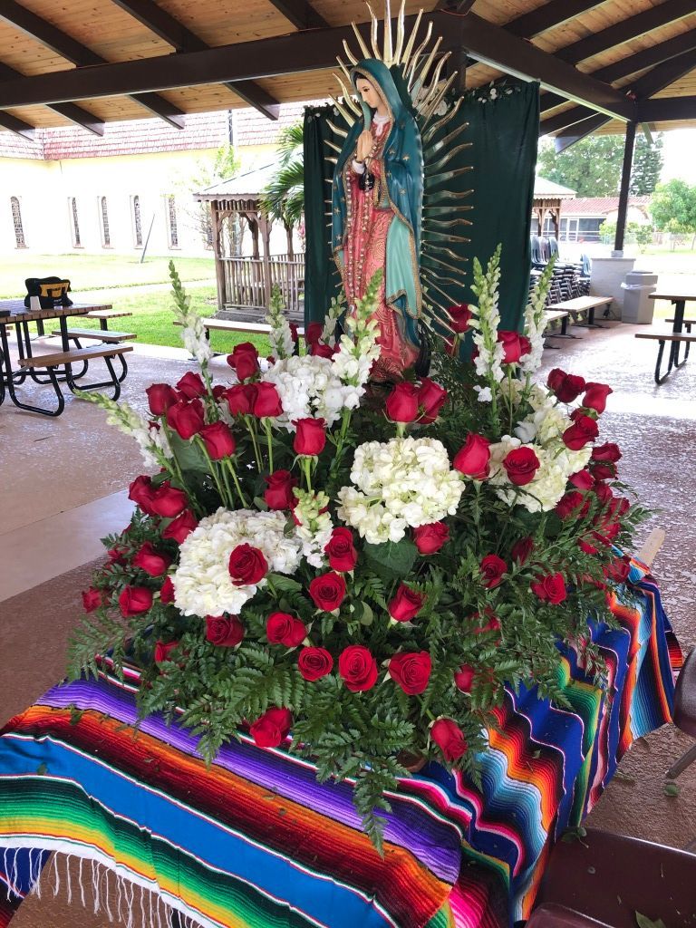 Floral altar with roses and white flowers beneath a Virgin Mary statue, covered in a colorful woven cloth.