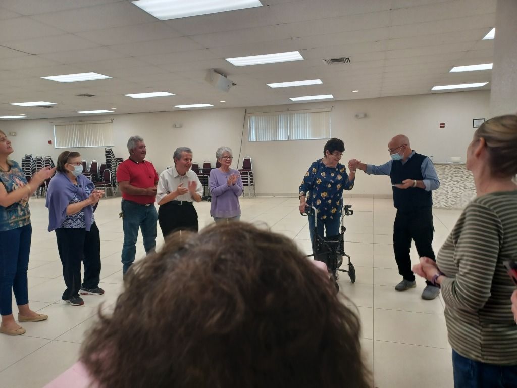 People dancing in a bright community hall, with others watching and clapping nearby.