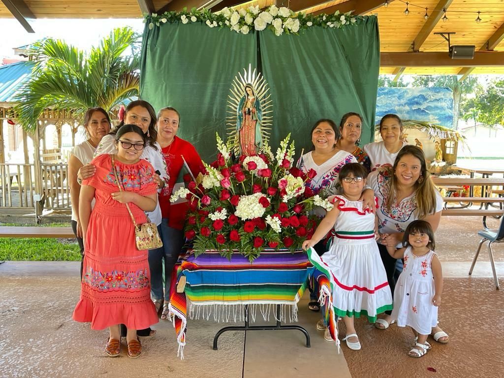 Group gathered beside a flower altar under a pavilion, with a green backdrop and smiling children in colorful dresses