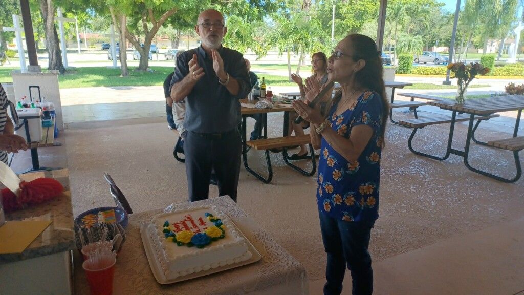 Two people stand by a birthday cake at a park pavilion, one clapping and one singing.