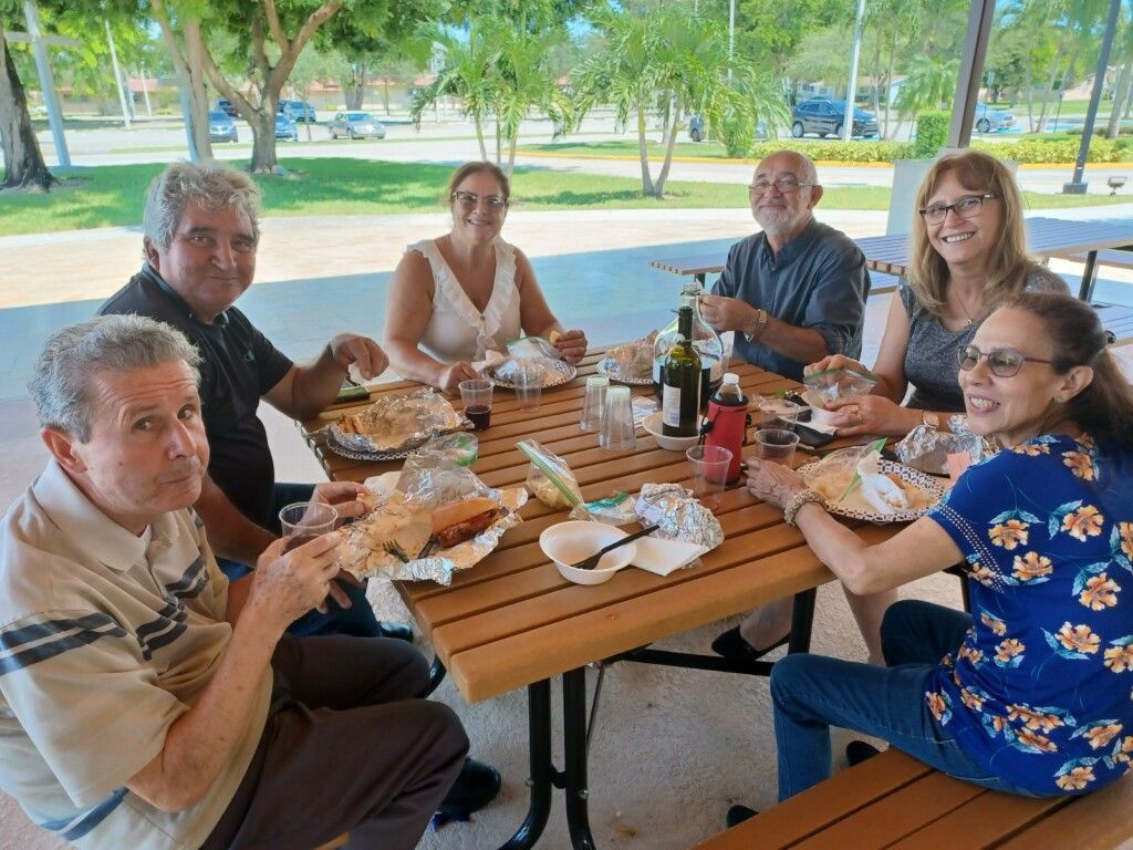 Six people share a picnic meal at a wooden table in a park pavilion.