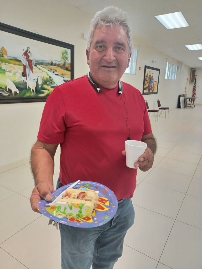Man holding a plate of food and a white cup in a bright hallway, smiling at the camera.