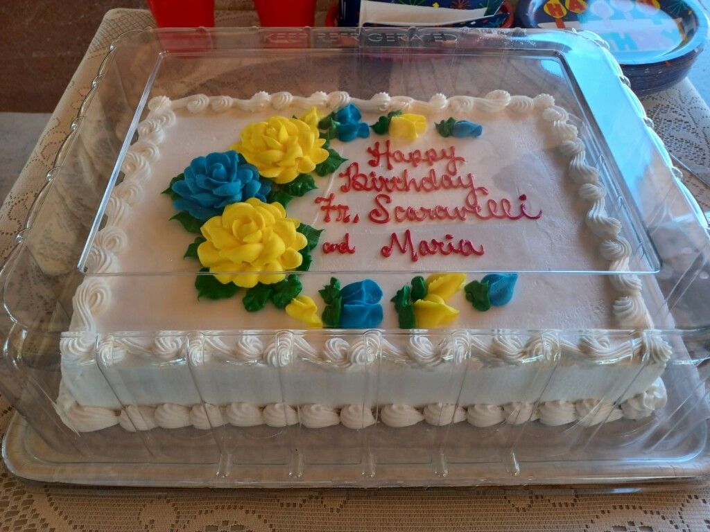 Decorated white cake with blue and yellow flowers and red Happy Birthday message in a clear box