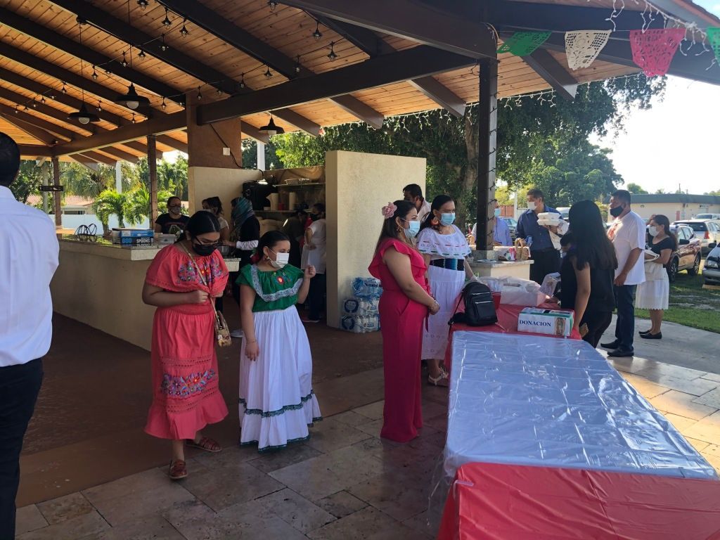 People standing under a pavilion beside a table with drinks, wearing face masks and dresses