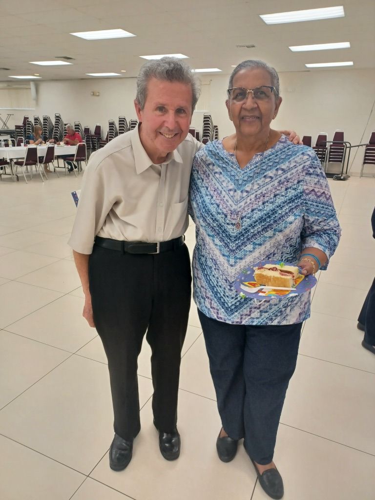 Two people smiling and standing together in a bright community hall, one holding a small item.
