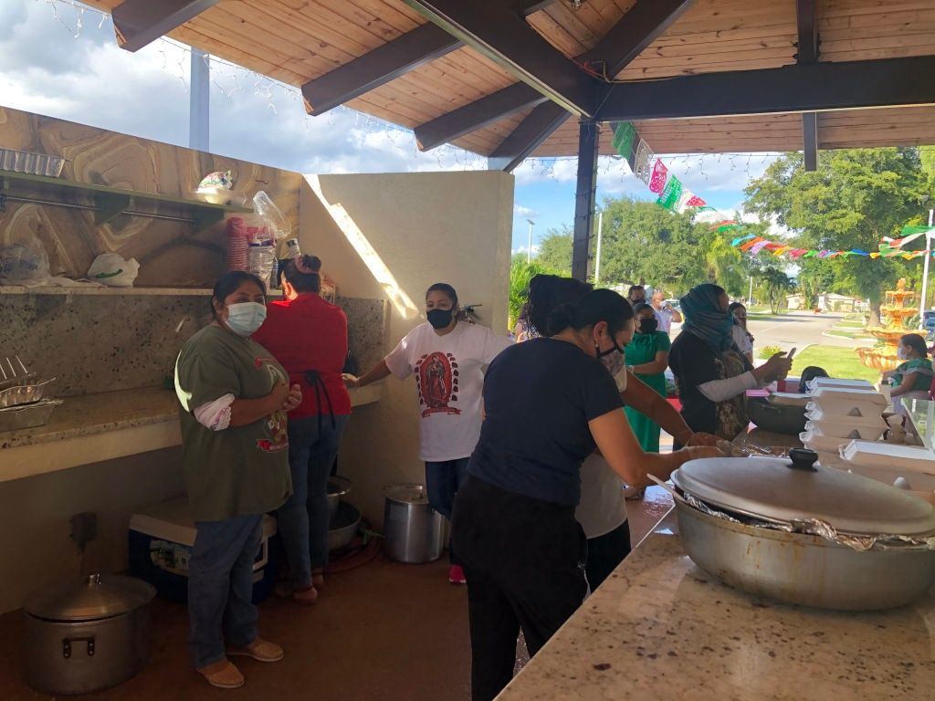 People serving food in a covered outdoor kitchen with large pots and trays