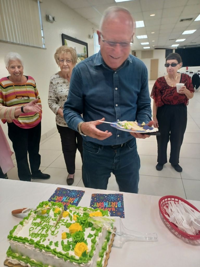 Man holding a plate with cake slices beside a decorated sheet cake at a small indoor celebration