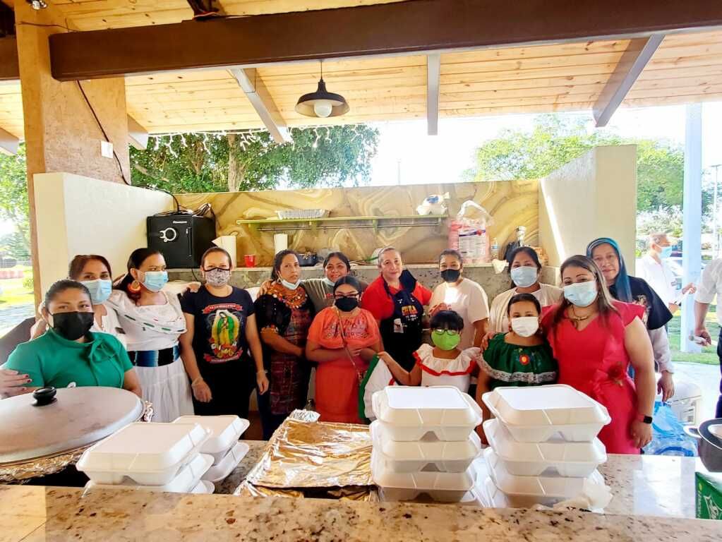 Group of people wearing masks gathered behind stacked takeout containers at a covered outdoor serving area.