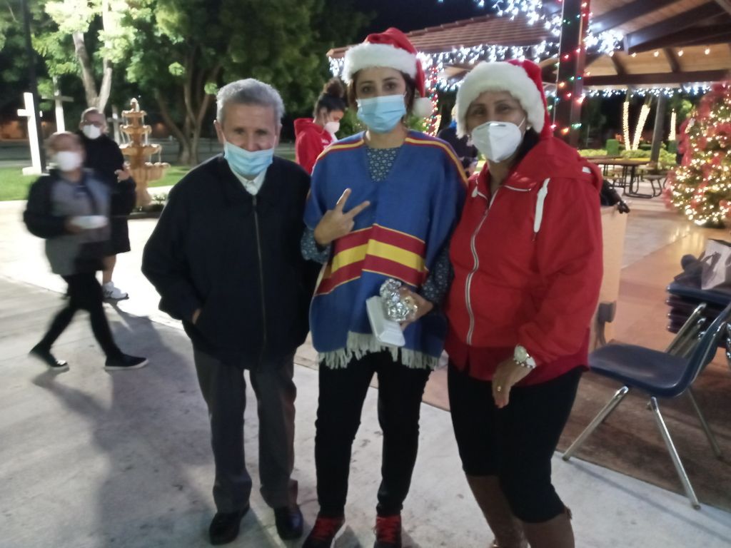 Three people posing at a holiday-lit outdoor plaza, wearing masks and Santa hats.