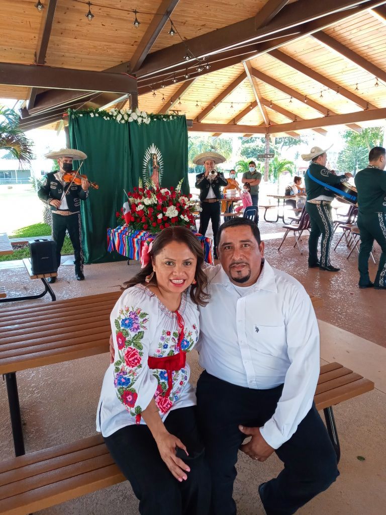 Couple seated on a bench in a pavilion, smiling with a floral display and musicians in the background