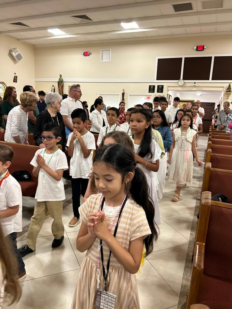 Children in white outfits stand in an auditorium aisle, some clasping hands in prayer during a gathering