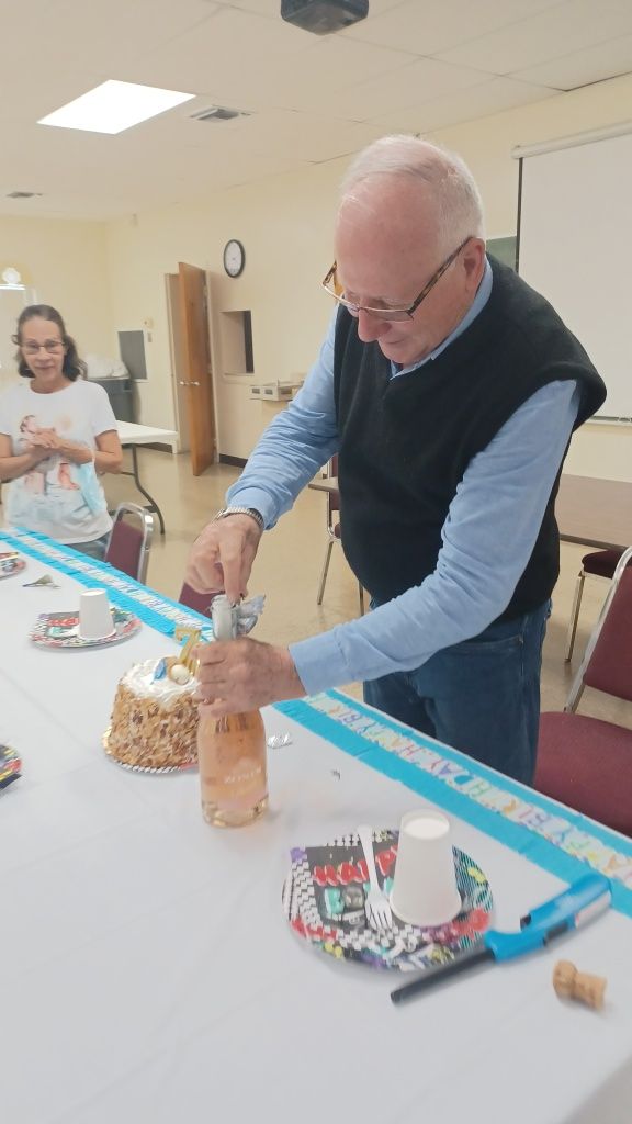 Man slicing a cake at a table in a community room, with snacks and drinks nearby.