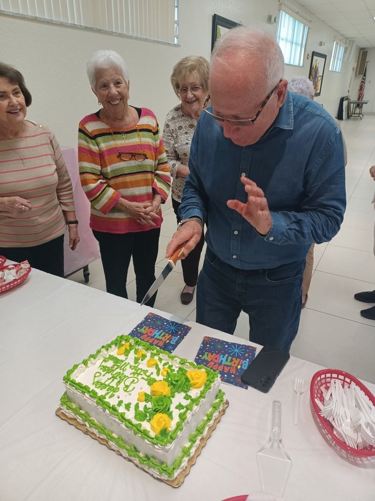 Man in blue shirt blowing out candles on a decorated birthday cake, with smiling people nearby.