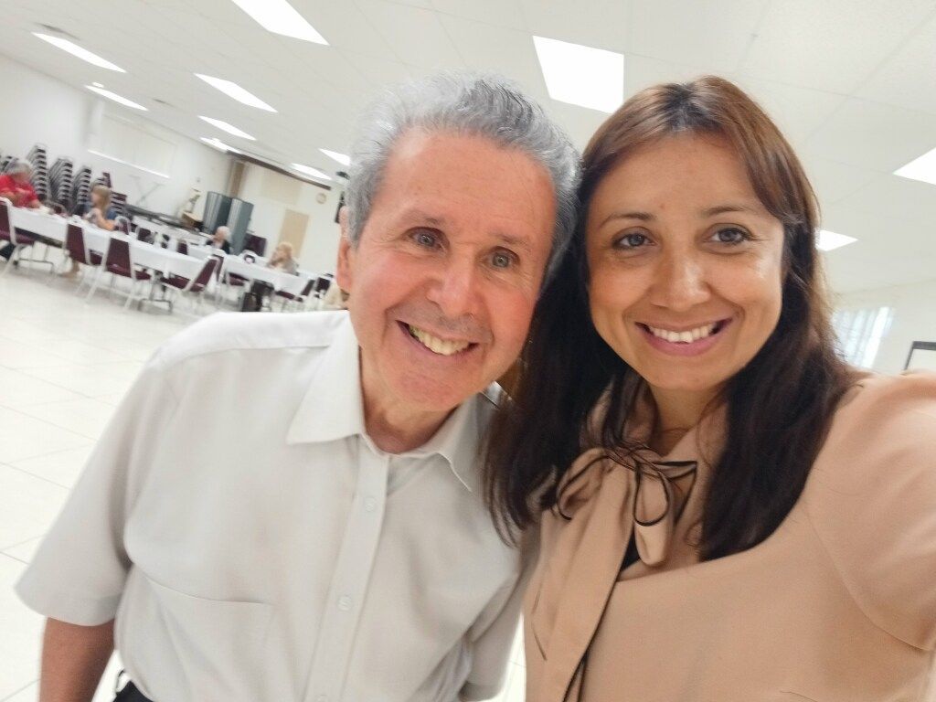 Two smiling people posing together in a bright room with tables and chairs in the background