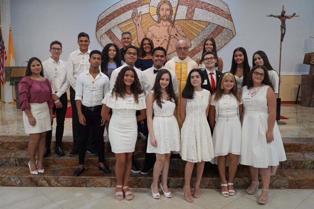 Group of people in white attire posing in a church before a large religious mural and crucifix.