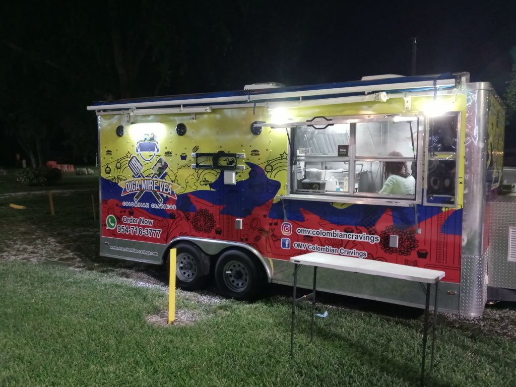 Night food truck with bright yellow and red wrap, lit serving window, parked on grass at dusk
