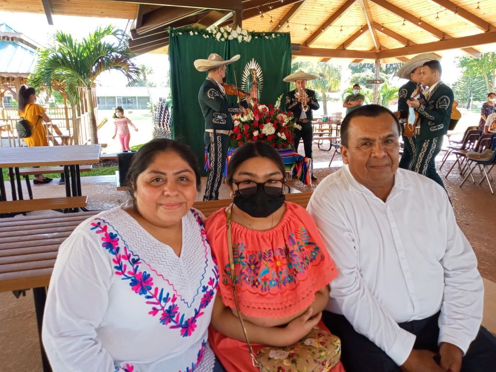 Three people seated at an outdoor pavilion with performers and a mariachi band in the background