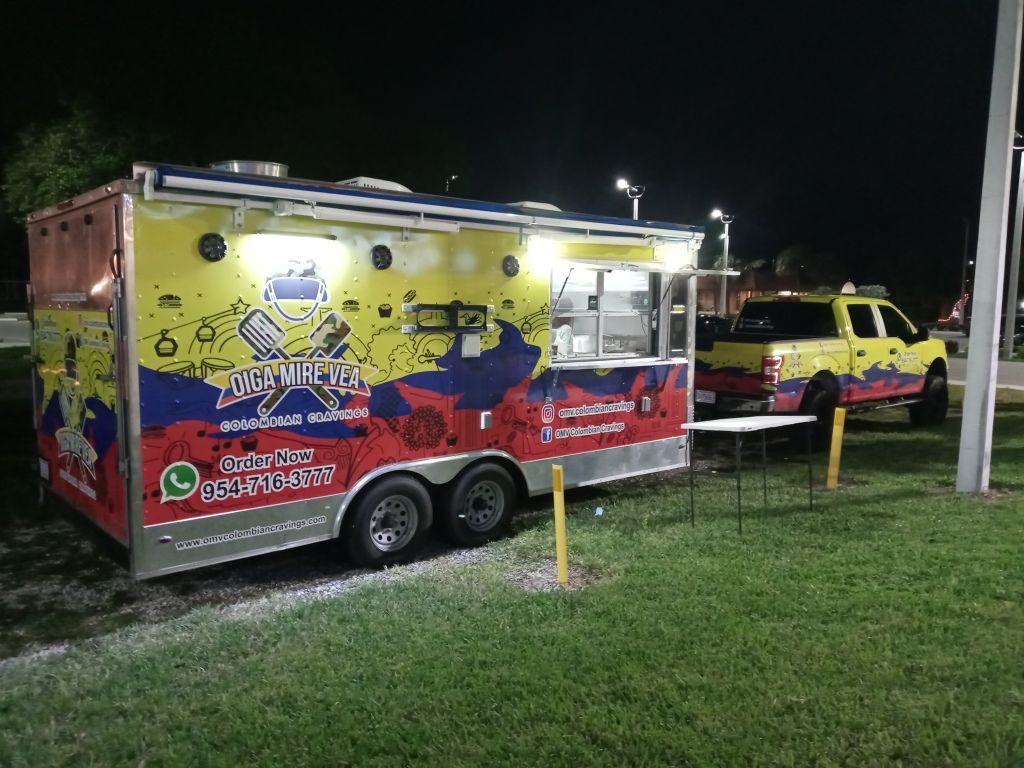 Bright yellow and red food truck with open serving window parked at night beside a yellow pickup truck.