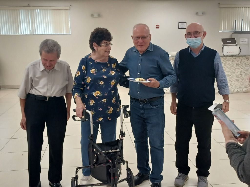 Four people stand indoors; one uses a walker while another receives a plate from a masked man.
