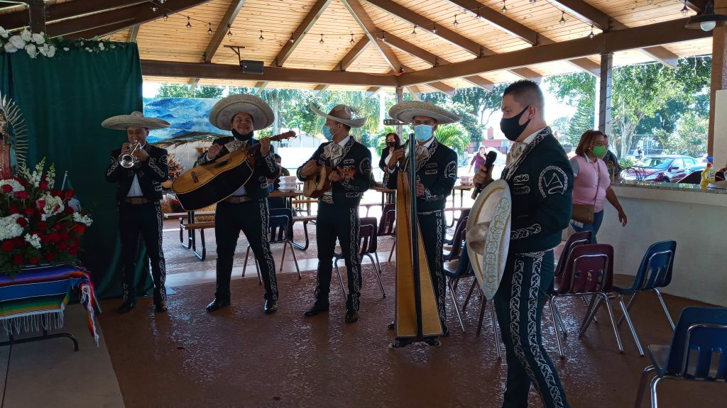 Musicians performing under a wooden pavilion, with microphones, drums, and chairs in a shaded outdoor setting