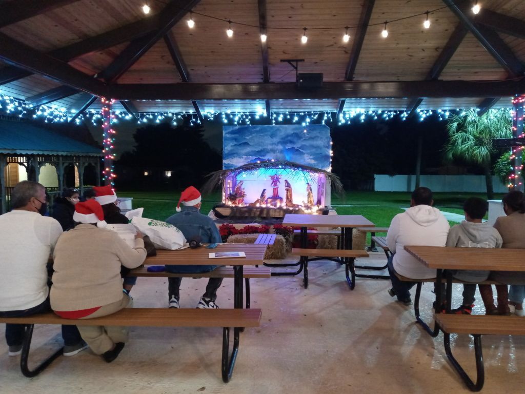 People seated on picnic benches under a pavilion watching a festive holiday stage performance.
