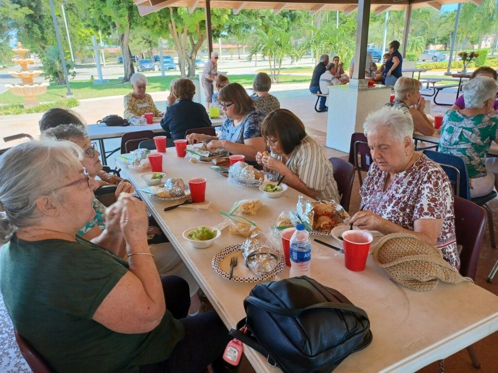 People eating at a long picnic table under a pavilion with drinks, plates, and baskets