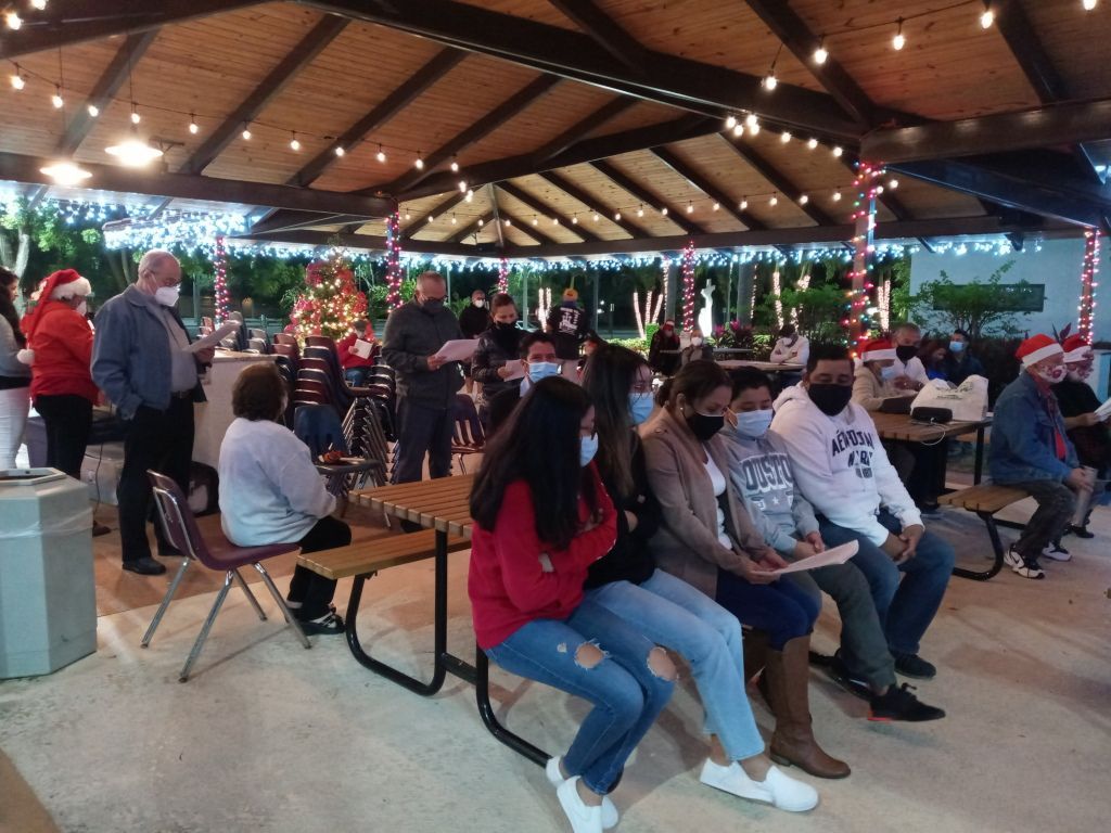 People seated under a pavilion at an outdoor gathering, with string lights and wooden beams overhead.