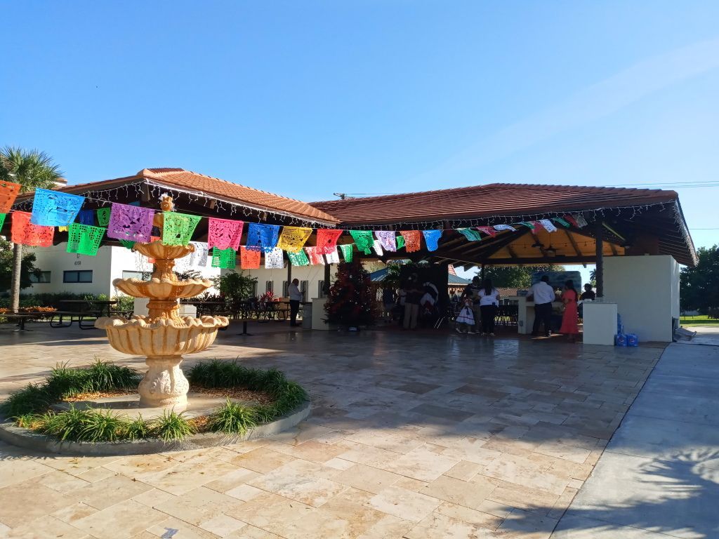 Outdoor plaza with colorful bunting, a fountain, and a covered seating area under a blue sky
