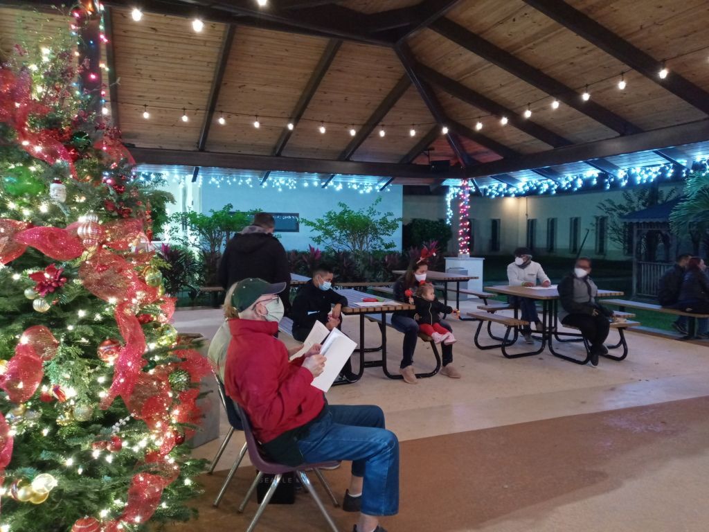 People seated under a lit pavilion beside a decorated Christmas tree and holiday lights.