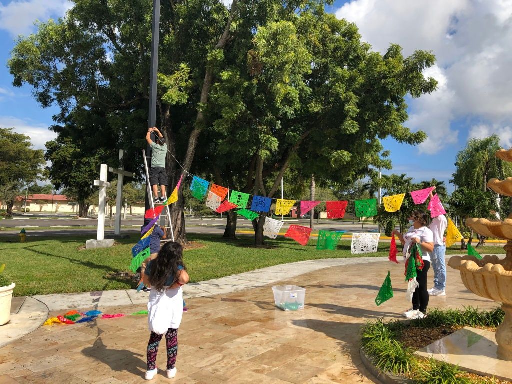 Children hanging colorful paper flags on a line in a sunny park with trees and benches