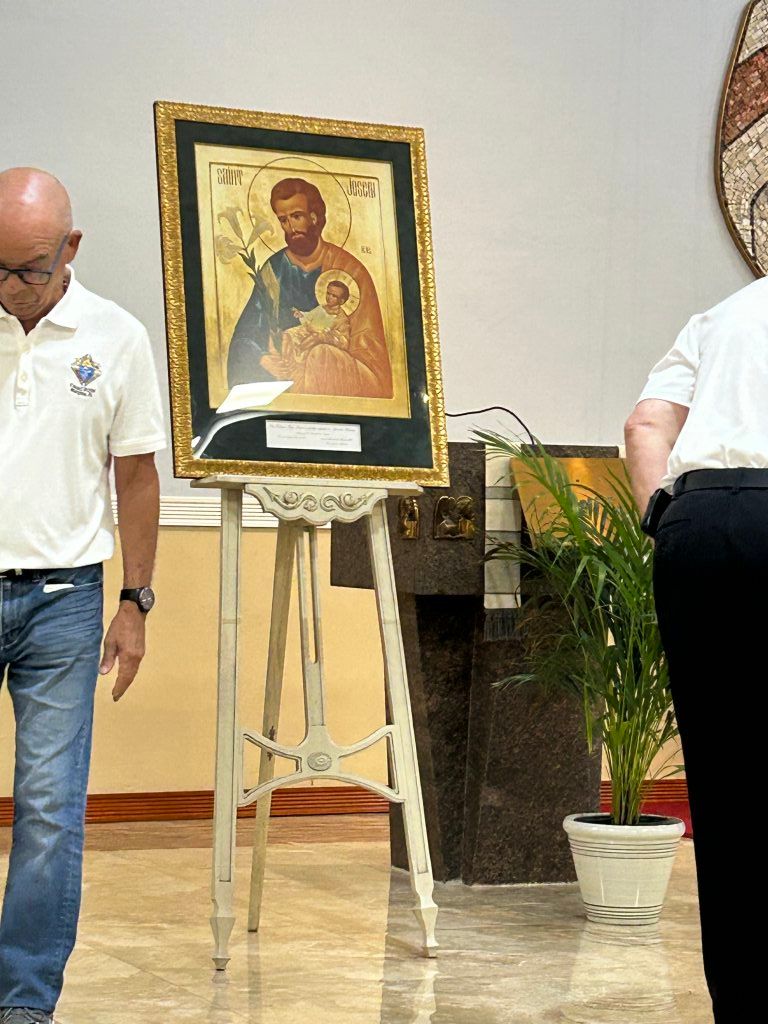Framed religious icon on an easel in a room, with two people partially visible on either side.