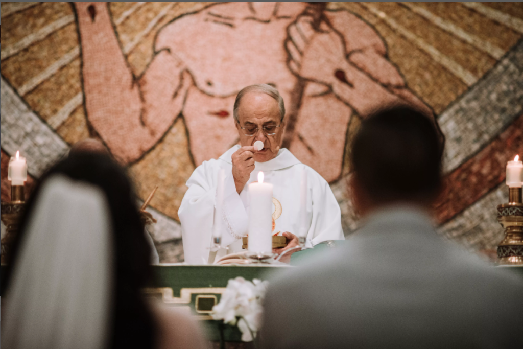 Priest at altar during church service, seen past seated congregation, with candles and a religious mural behind him