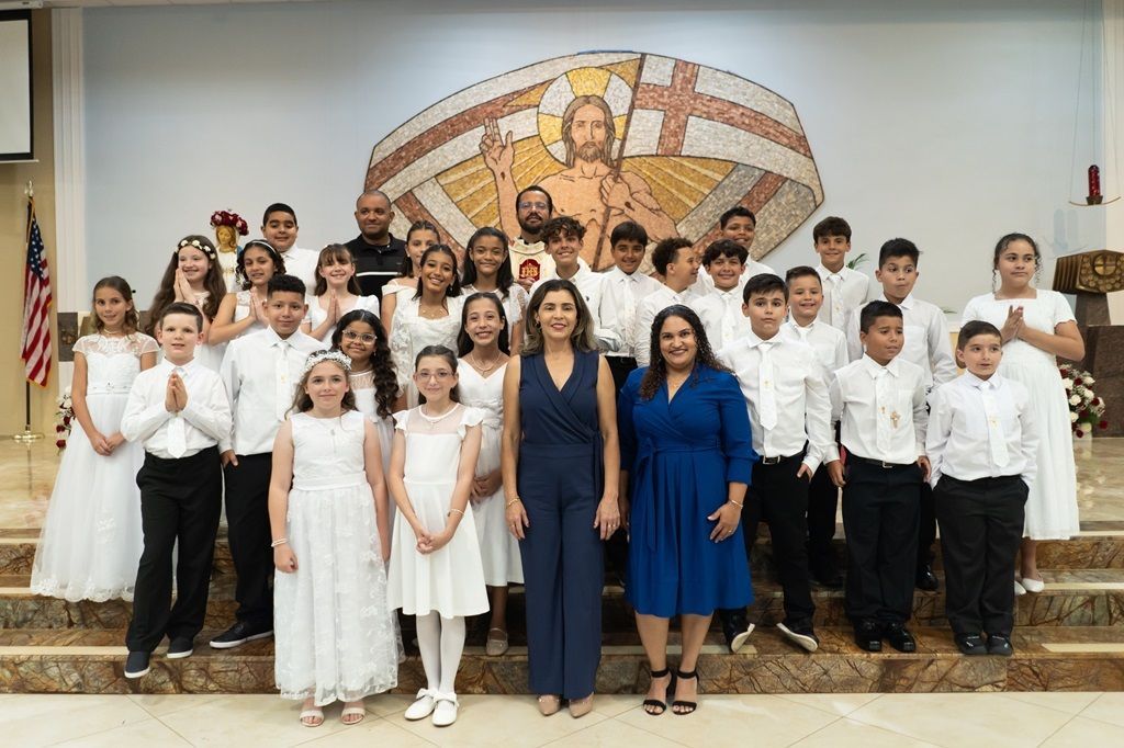 Group of children in white outfits posing on church steps in front of a mosaic wall.