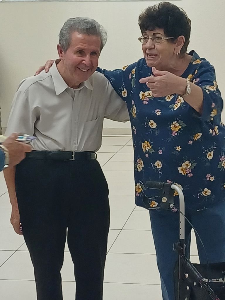 Two people smiling and posing together indoors, one with an arm around the other, beside a walker