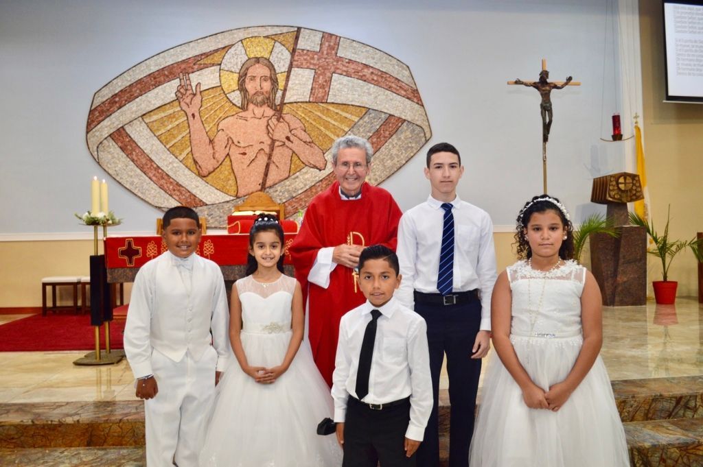 Children and a priest stand in a church in front of a mural and crucifix, dressed in white and formal wear