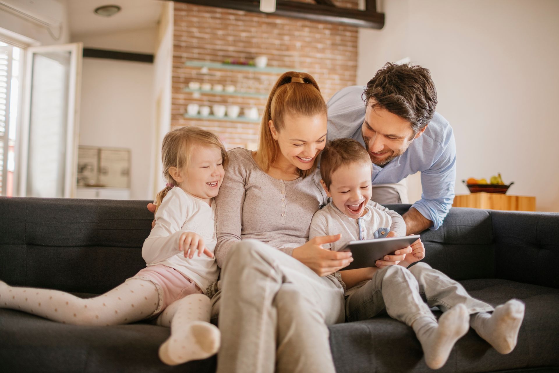 Family of four laughing while looking at a tablet together on a sofa in a living room.