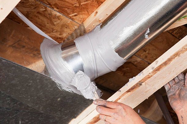 Ductwork being sealed with gray sealant and tape in a wood-framed ceiling. Hands are visible.