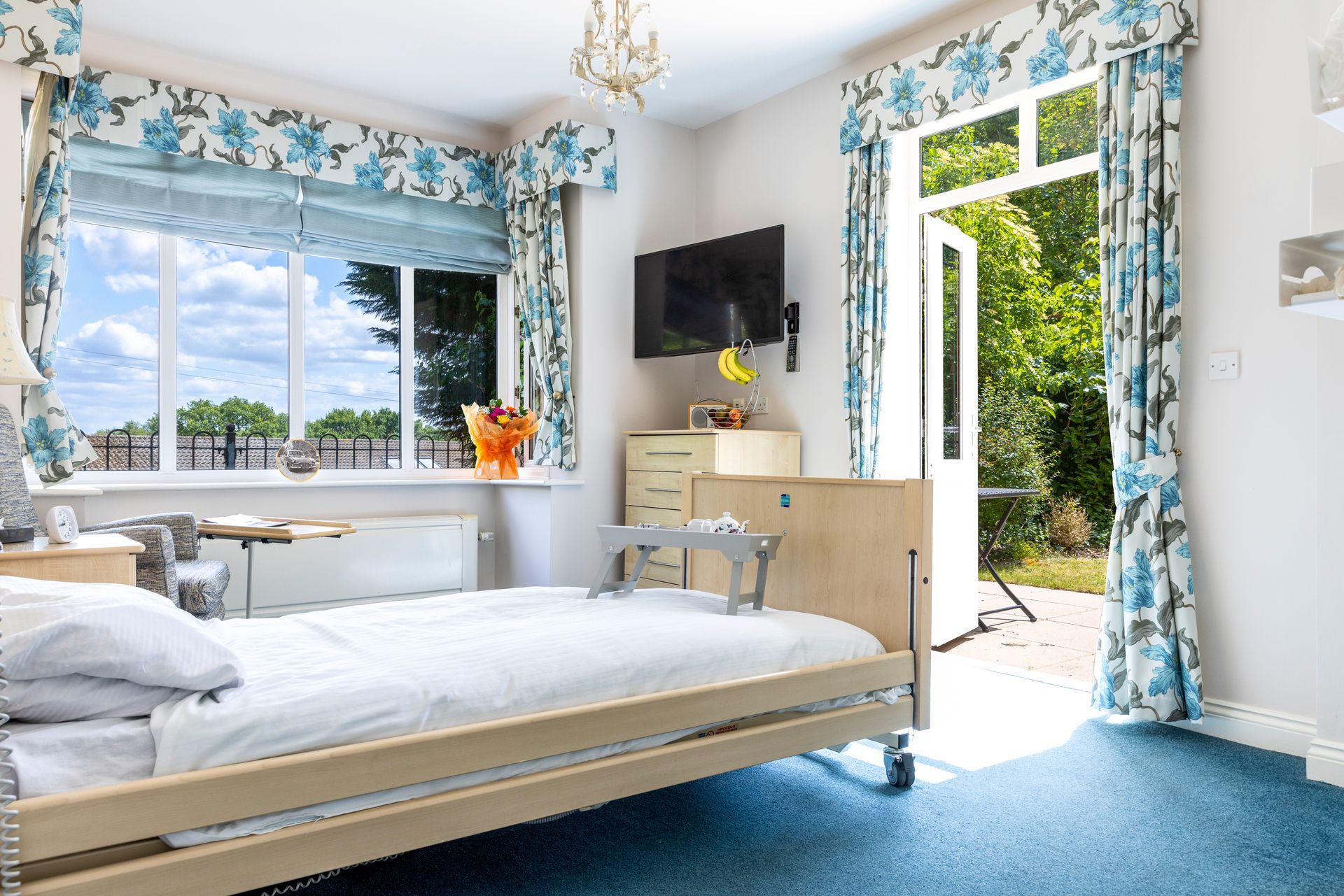 Bedroom with a hospital bed, window with a view, and open doors to the outside. Floral curtains and blue carpet.