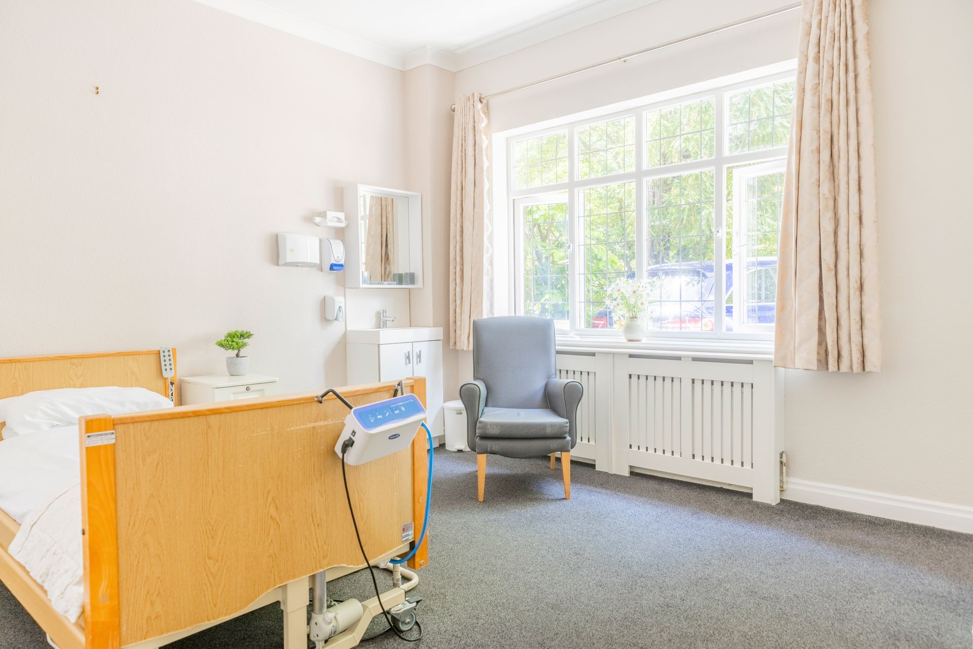 Bedroom with a hospital bed, armchair, and large window with light-colored curtains. There is a sink and medicine cabinet.