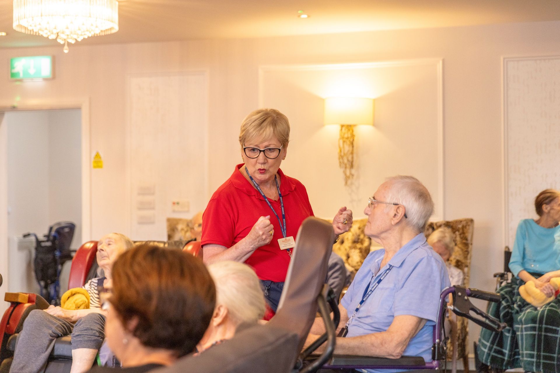 Woman in red shirt gestures to seated senior in a blue shirt. They are inside a room with other seated seniors.