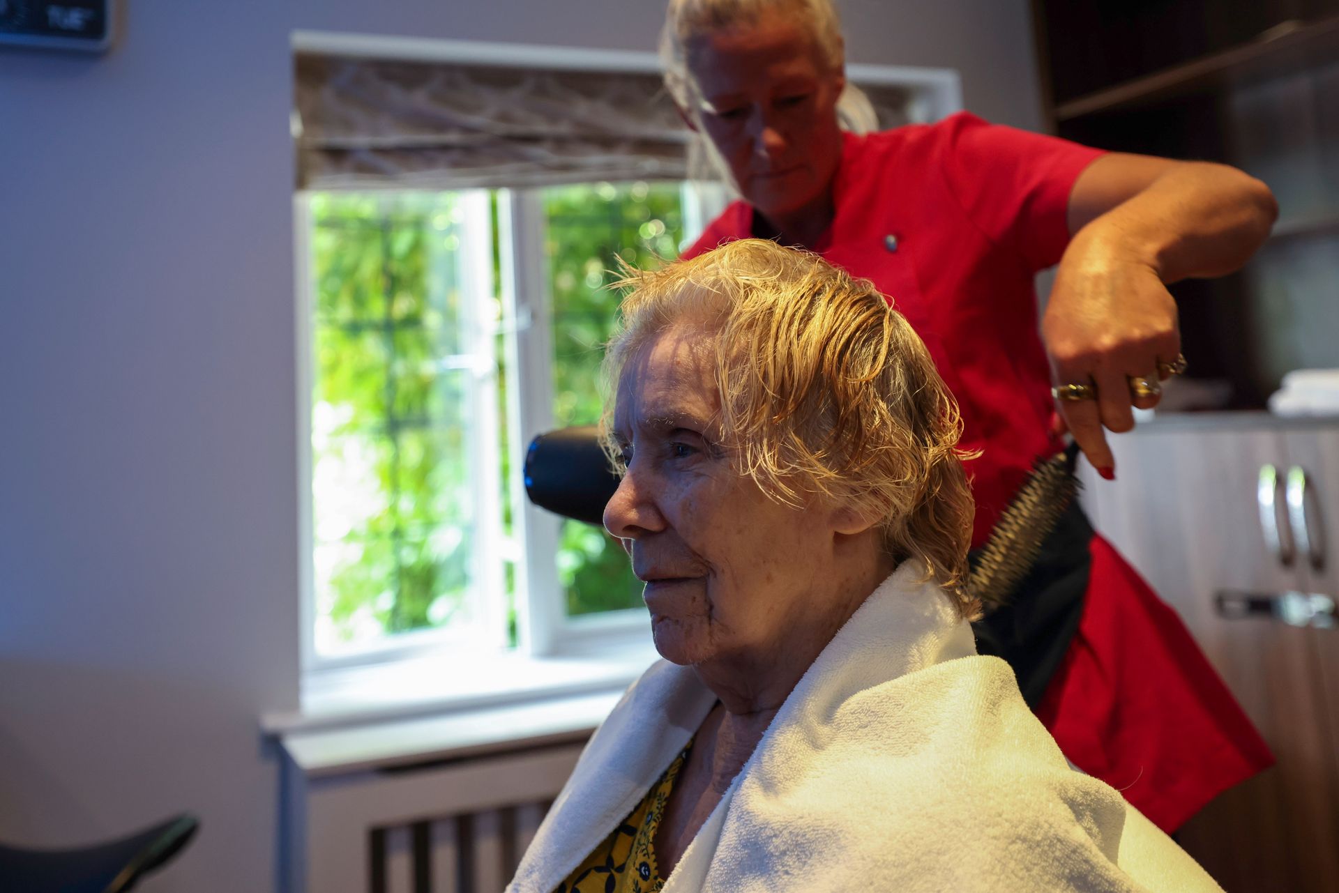 A woman in a white towel gets her hair cut by a person in a red shirt near a window.