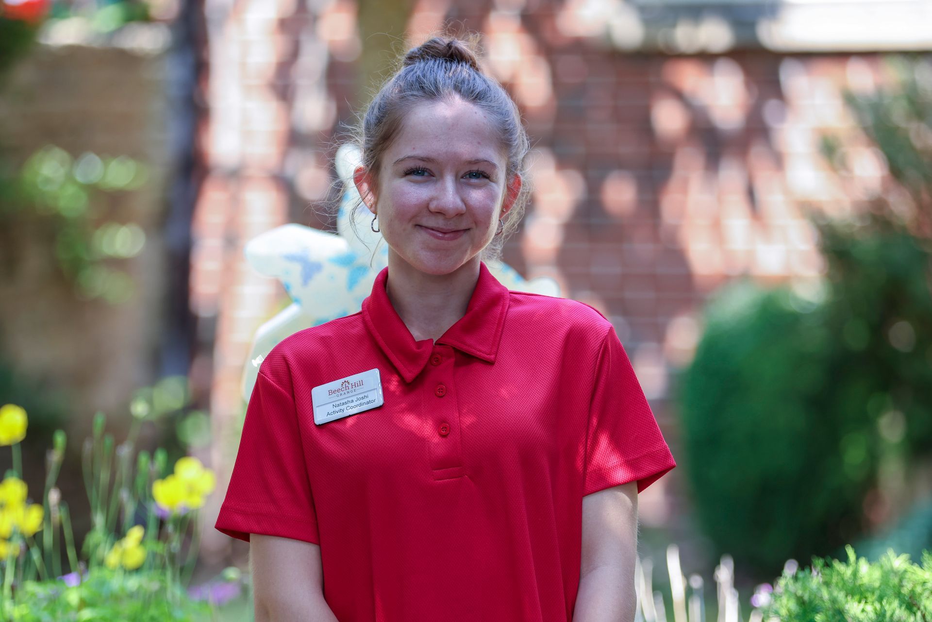 Young woman with a bun smiles while wearing a red polo shirt and name tag, standing in a garden.