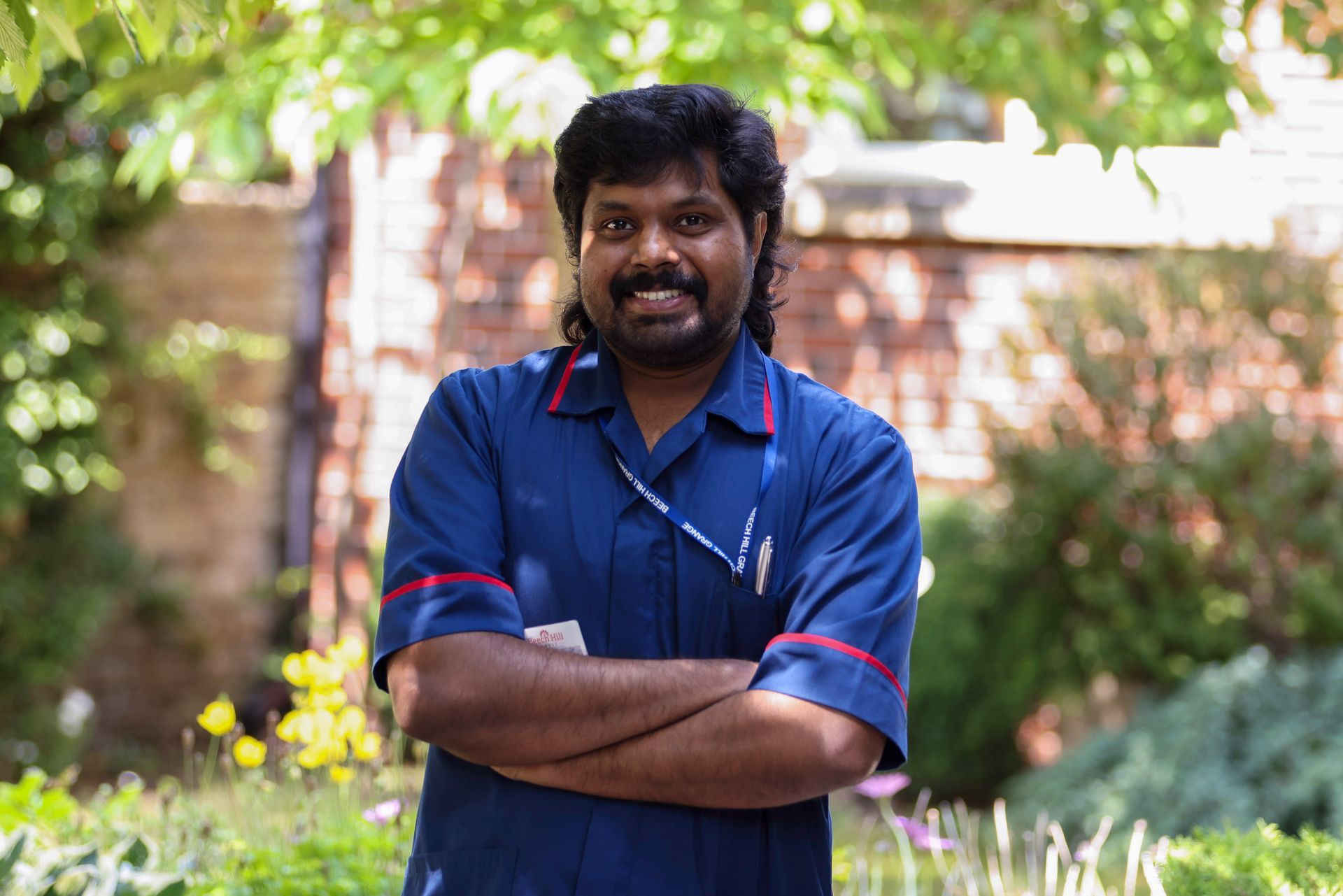 Man with dark hair and a mustache wearing a blue uniform with crossed arms, standing outdoors in front of a garden.
