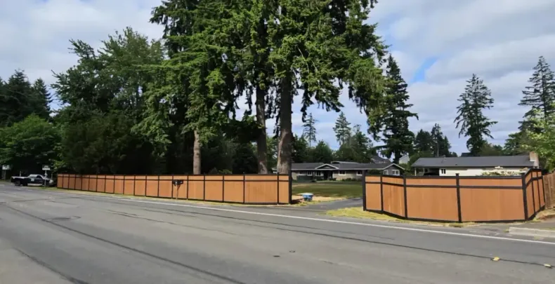 Wooden fence lining a roadside yard with large trees and a house in the background