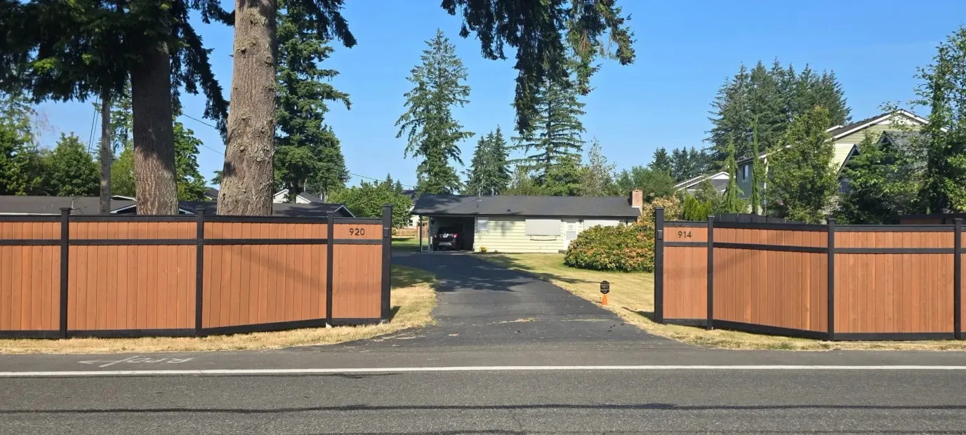 Open wooden gate with a driveway leading to a house, framed by tall trees under a blue sky
