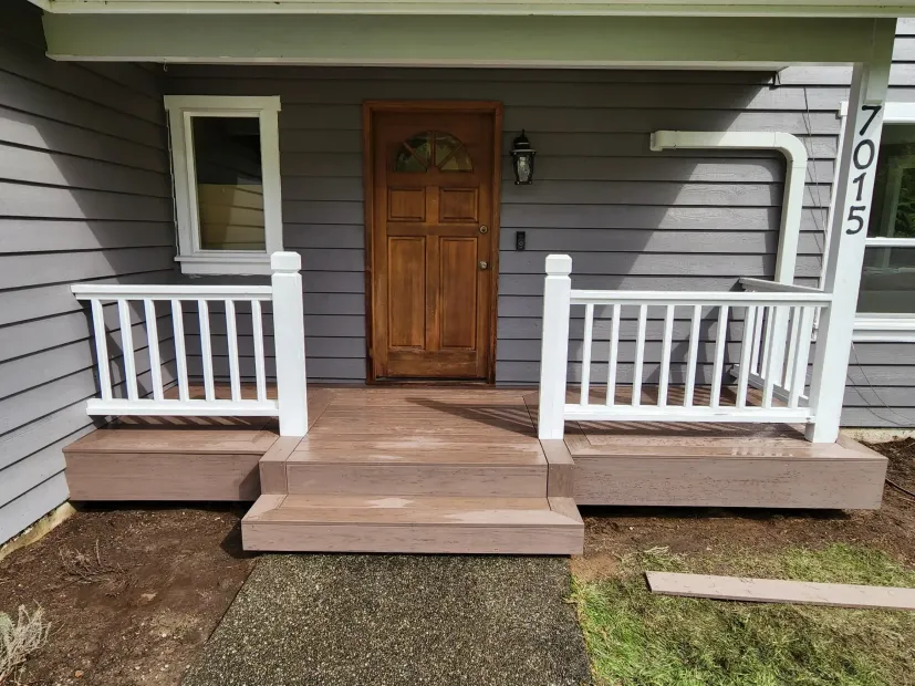 Front porch with wooden steps, white railings, and a brown door on gray siding.