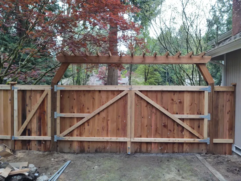 Wooden backyard gate and fence with diagonal braces beneath a pergola frame, trees in the background